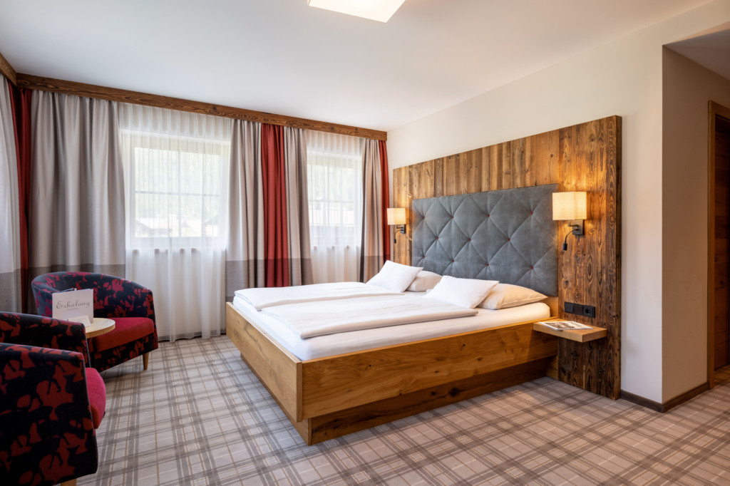 Helles Hotelzimmer mit Holzbett, gepolstertem Kopfteil, Sesseln und großen Fenstern
